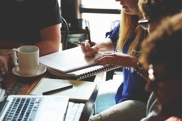 Personas colaborando en una mesa con un ordenador portátil, un cuaderno y café en una oficina luminosa.