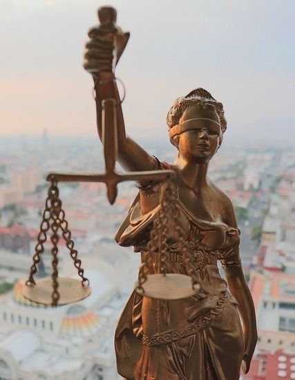 Estatua de bronce de la Dama de la Justicia sosteniendo una balanza, con un paisaje urbano borroso al fondo.