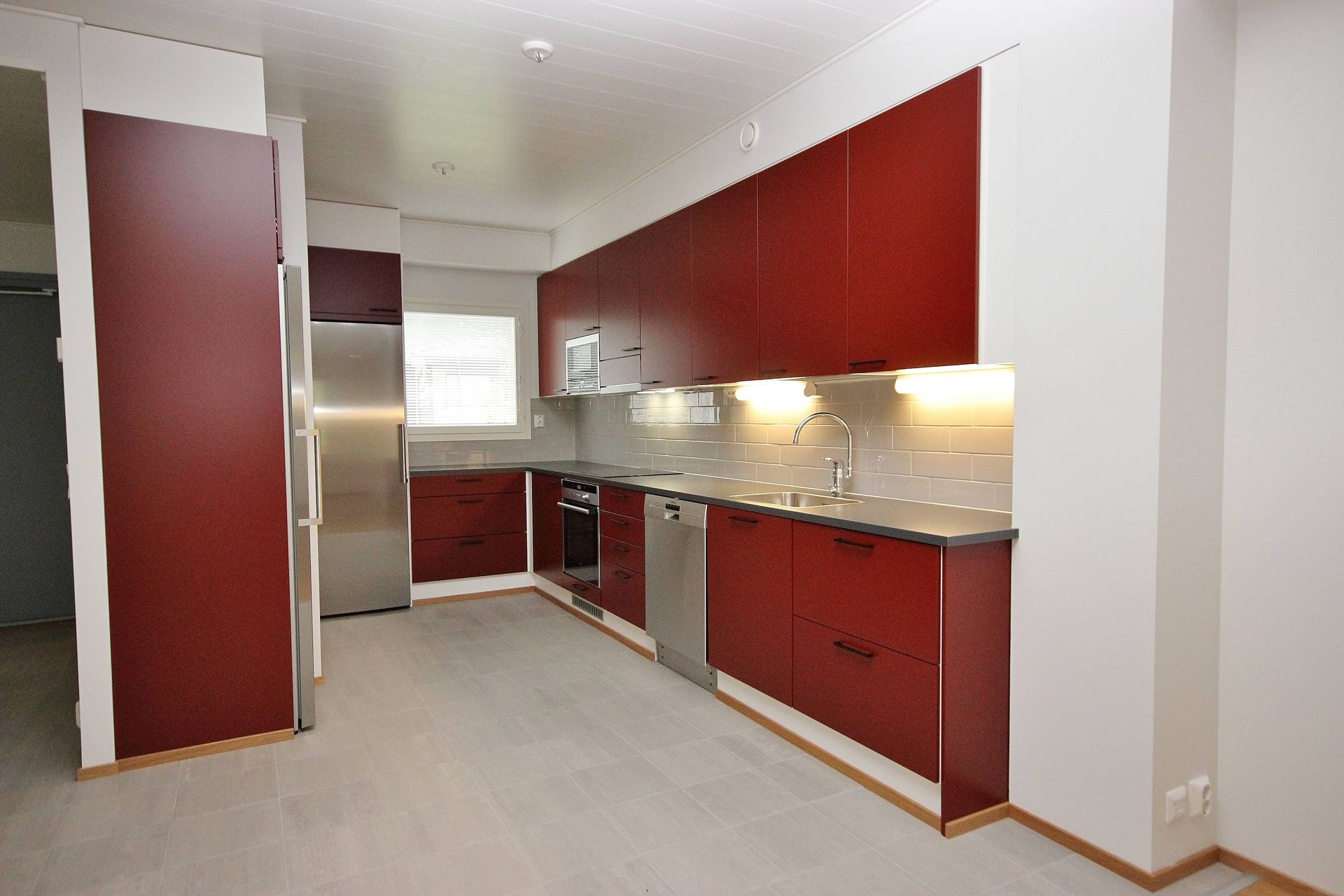 Moderni keittiö, jossa punaiset kaapit, ruostumattomasta teräksestä valmistetut kodinkoneet ja vaaleat työtasot
