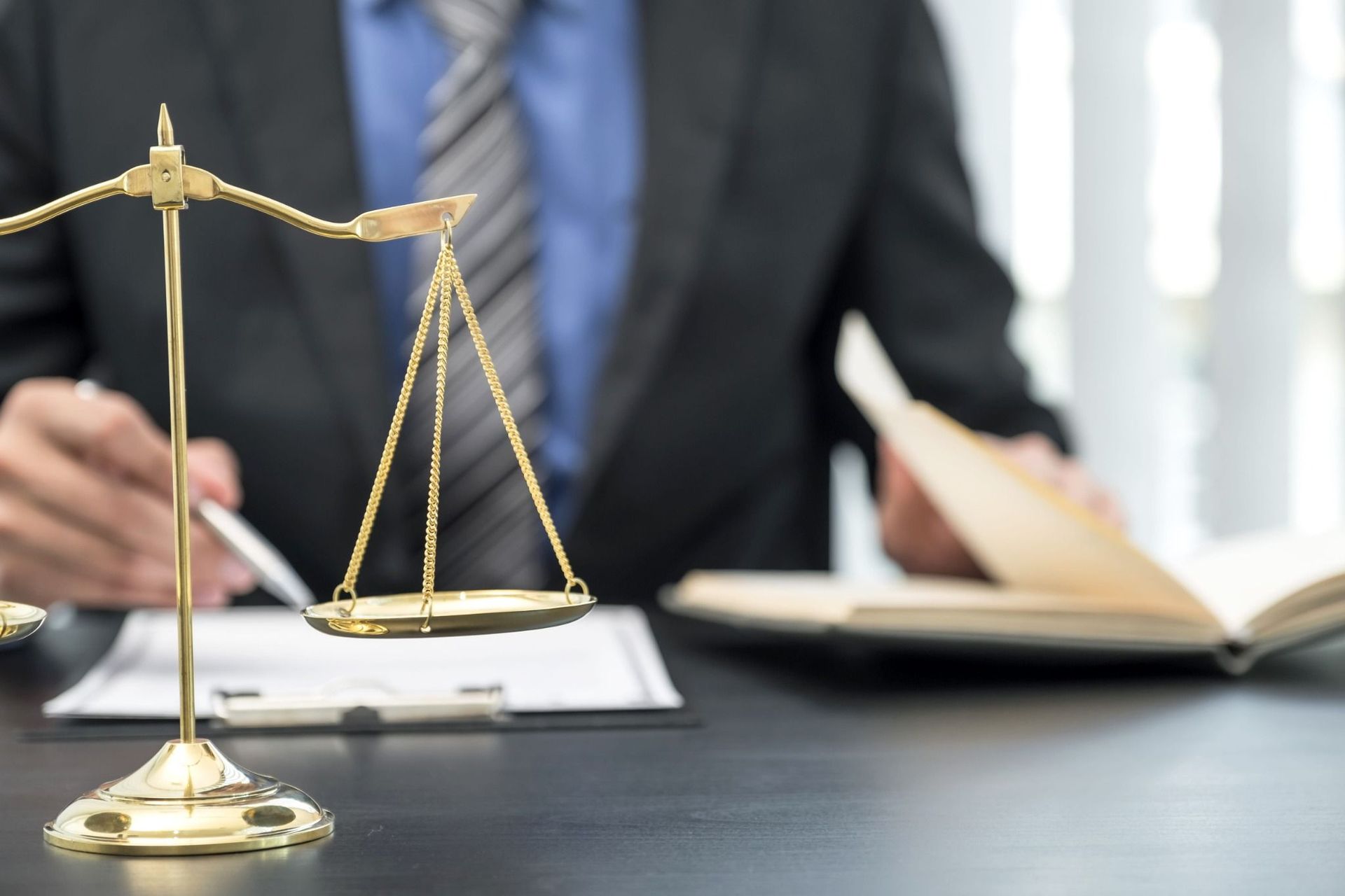 Balanza dorada sobre un escritorio con una persona vestida de traje escribiendo al fondo, simbolizando la ley o la justicia.
