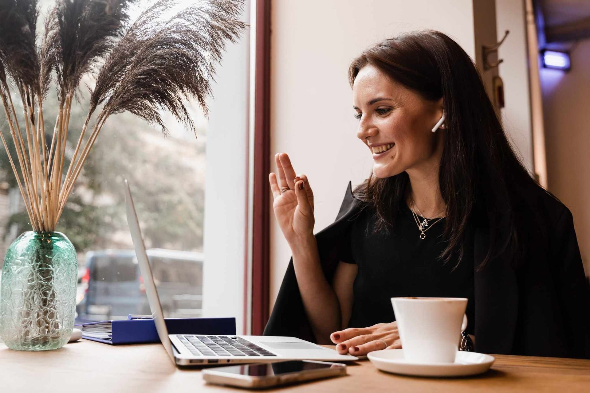 Una mujer sentada en la mesa de un café, sonriendo y saludando durante una videollamada por portátil.