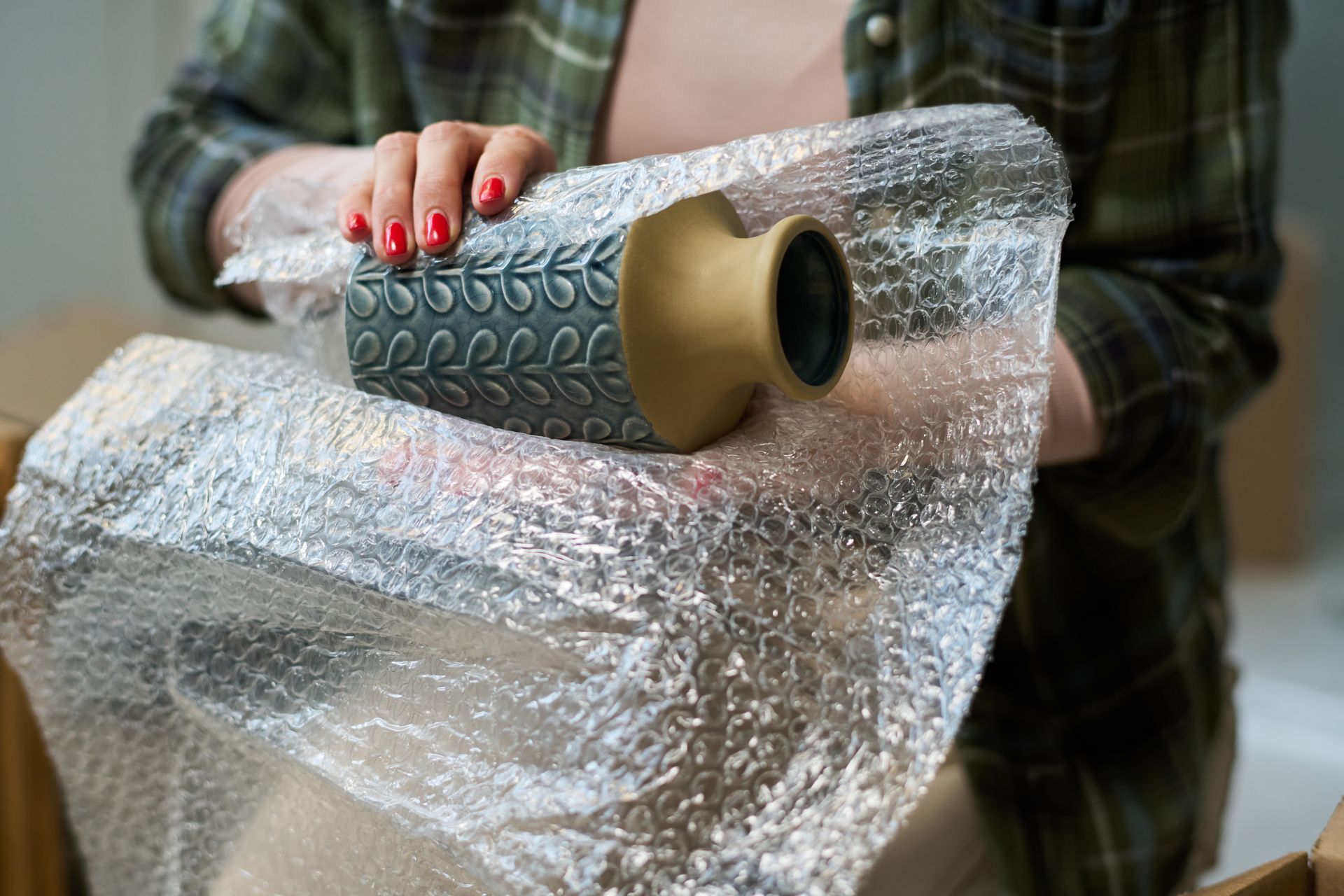 Une personne portant une chemise à carreaux enveloppe un vase bicolore à motifs dans du papier bulle transparent.