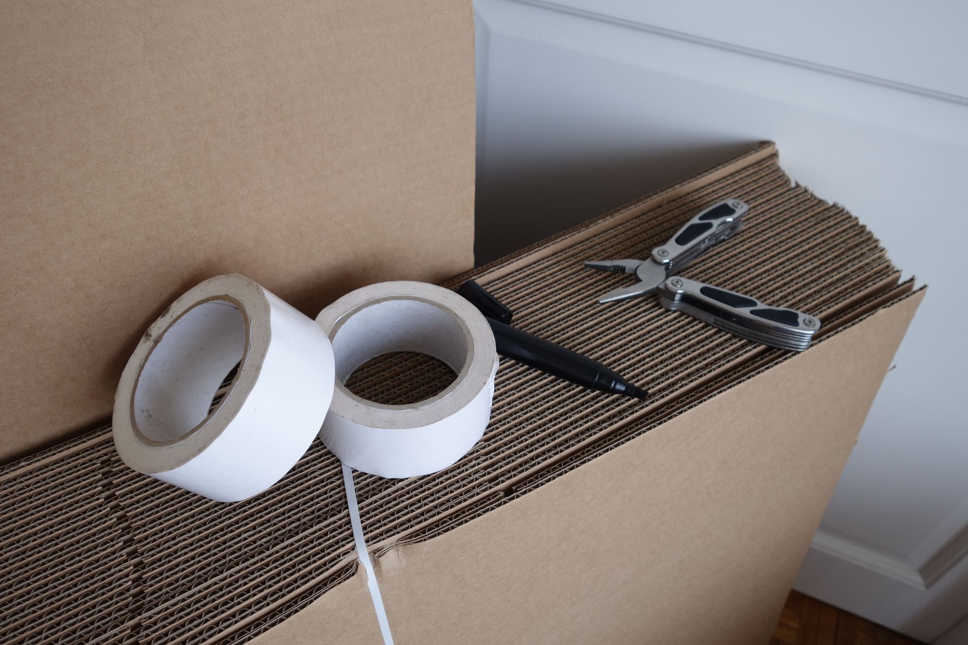 Deux rouleaux de ruban adhésif blanc, un stylo noir et un outil multifonction posés sur une pile de cartons à plat.