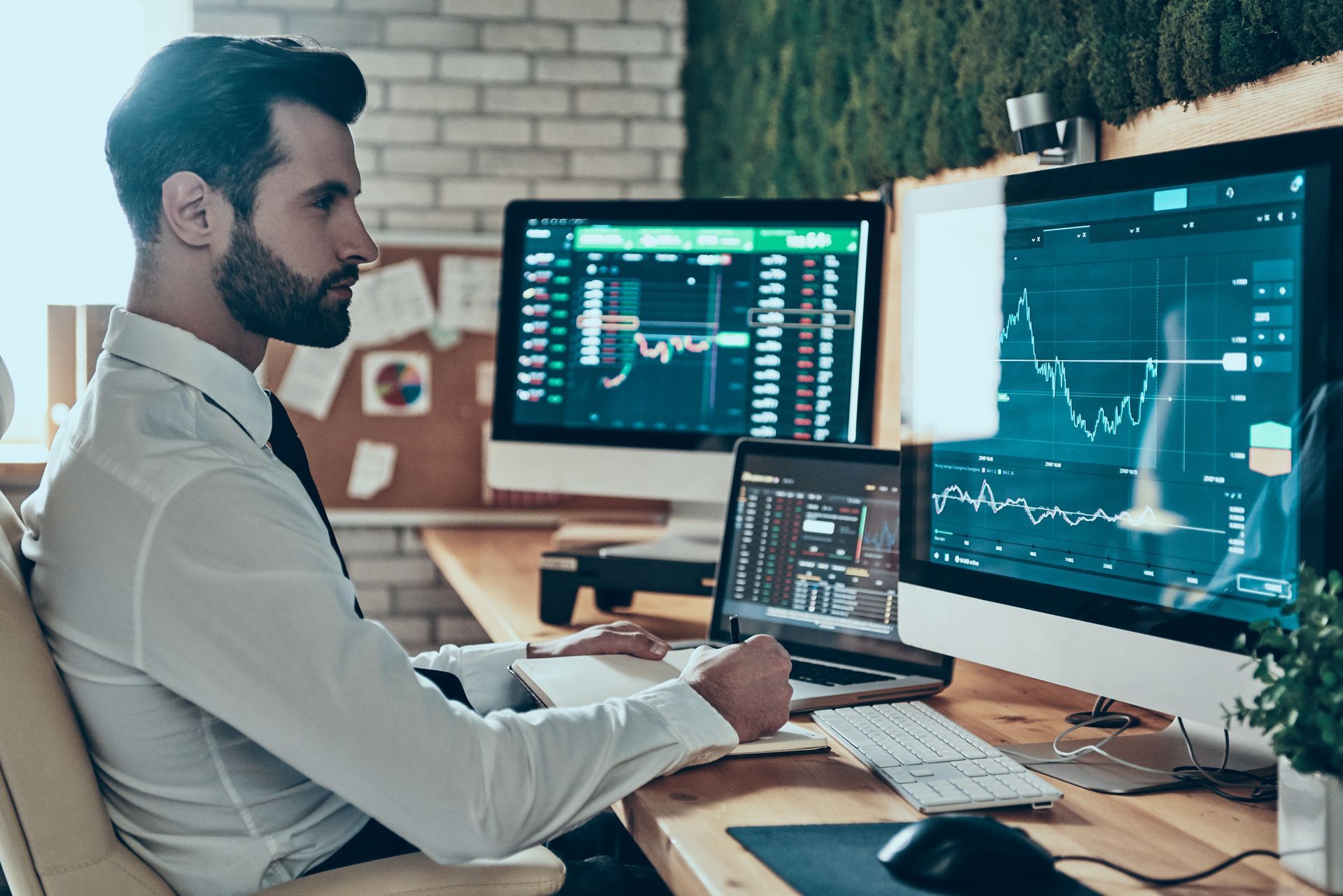 Une personne en chemise blanche écrit sur un carnet à un bureau où se trouvent plusieurs écrans affichant des graphiques boursiers.