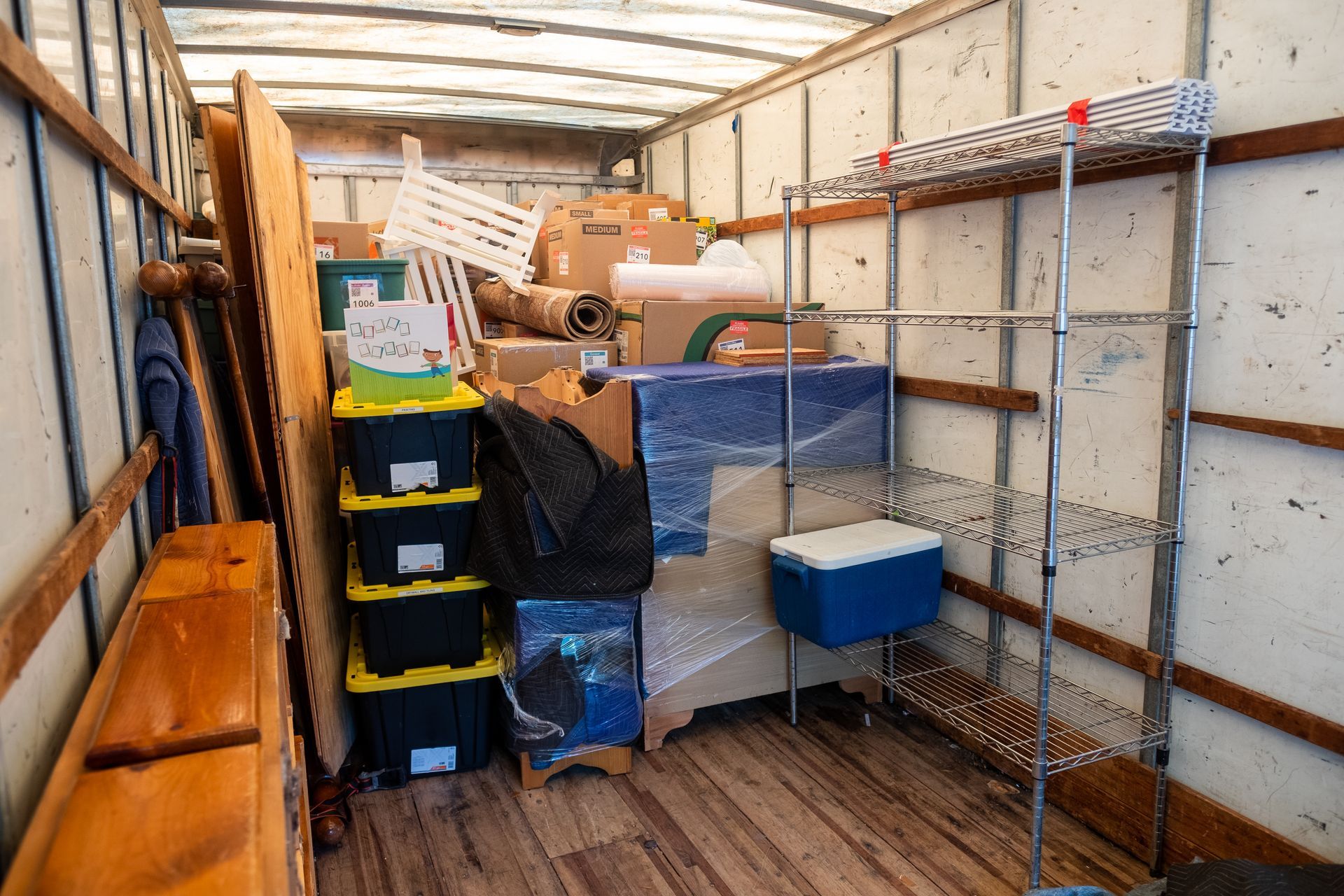 Dans un camion de déménagement, des meubles en bois, des bacs de rangement, une commode emballée et une étagère métallique sont entassés les uns sur les autres.
