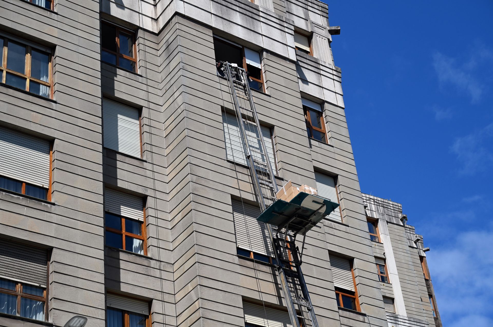 Une plateforme élévatrice de monte-meubles, levée contre la façade d'un immeuble, supporte un carton destiné à être introduit par une fenêtre.