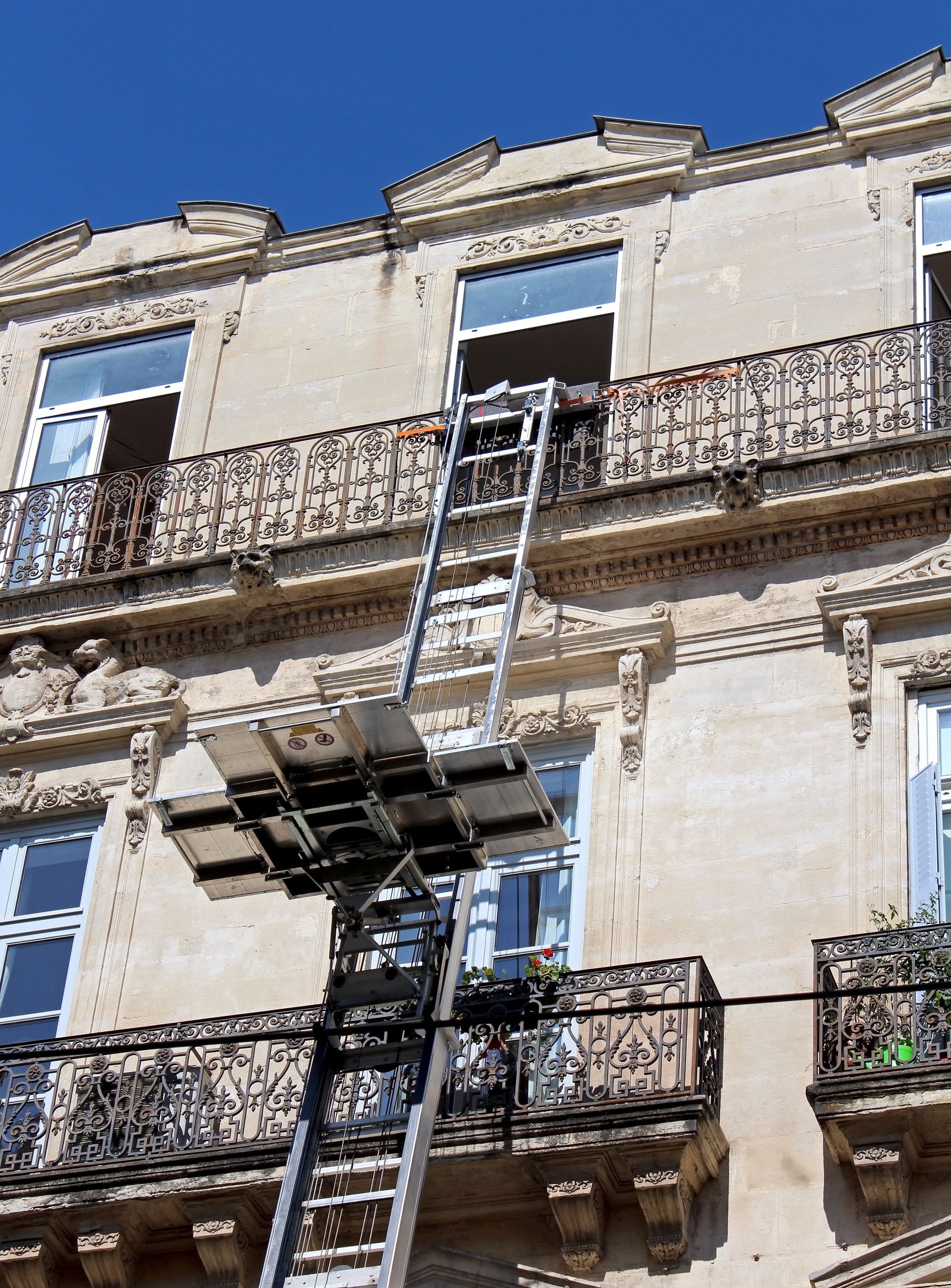 Un monte-meubles portatif permet d'accéder au balcon du deuxième étage d'un immeuble beige de style classique, sous un ciel bleu.