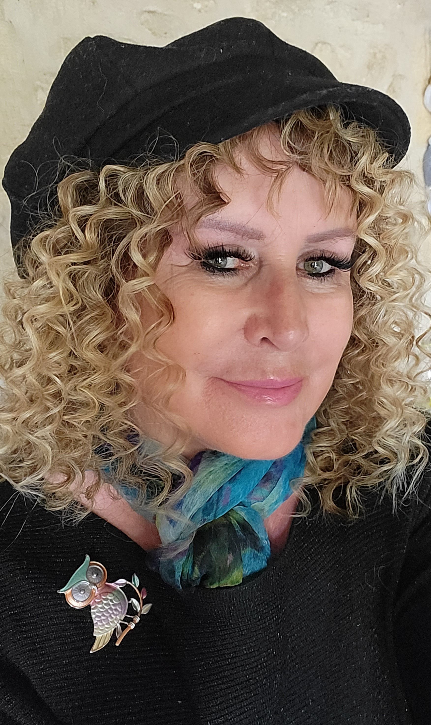 Femme aux cheveux blonds bouclés portant un chapeau noir, un pull noir et une écharpe bleue ; souriant à l'objectif.