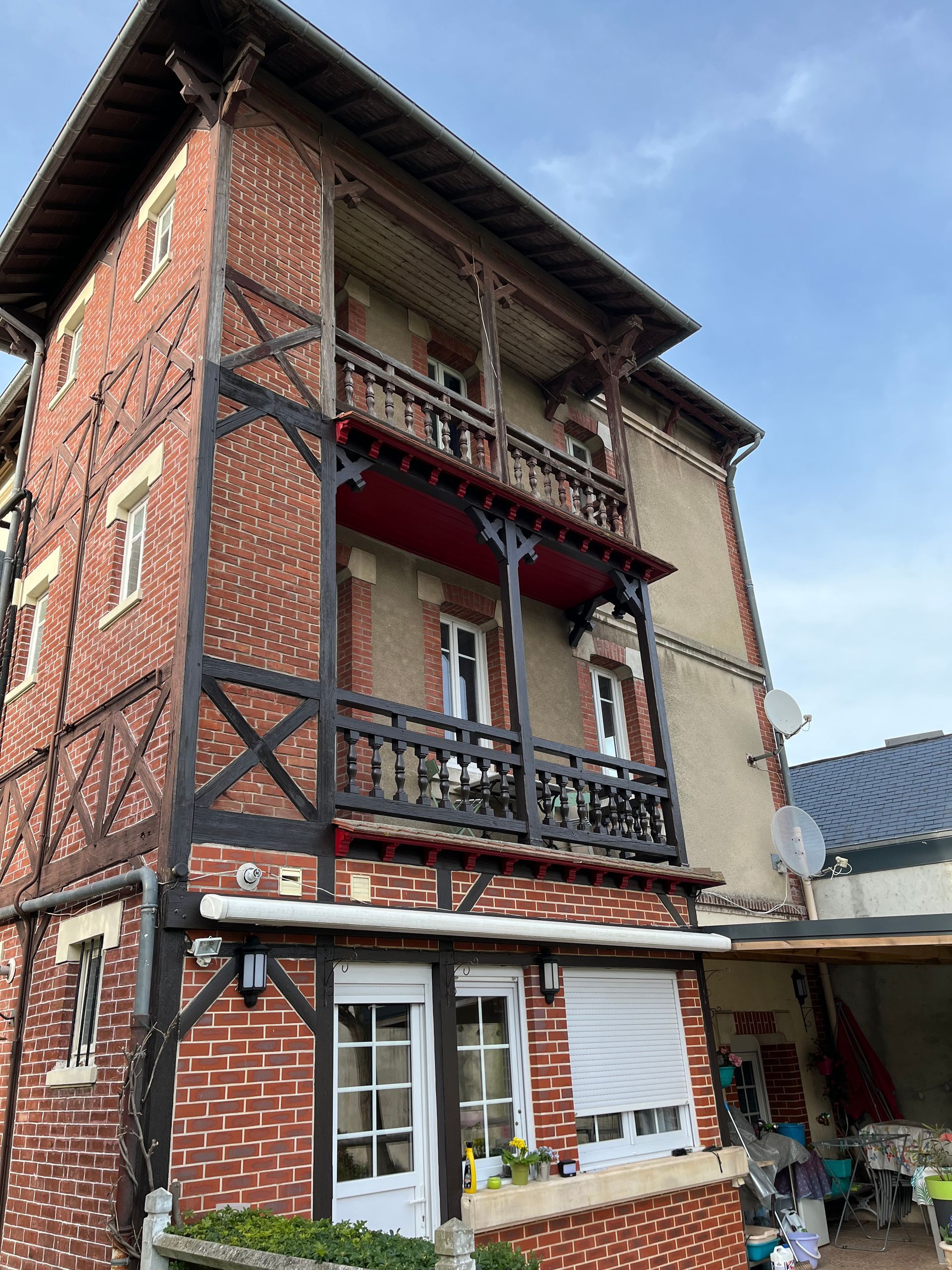 Maison en briques de trois étages avec balcons, volets rouges et porte blanche au rez-de-chaussée sous un ciel nuageux