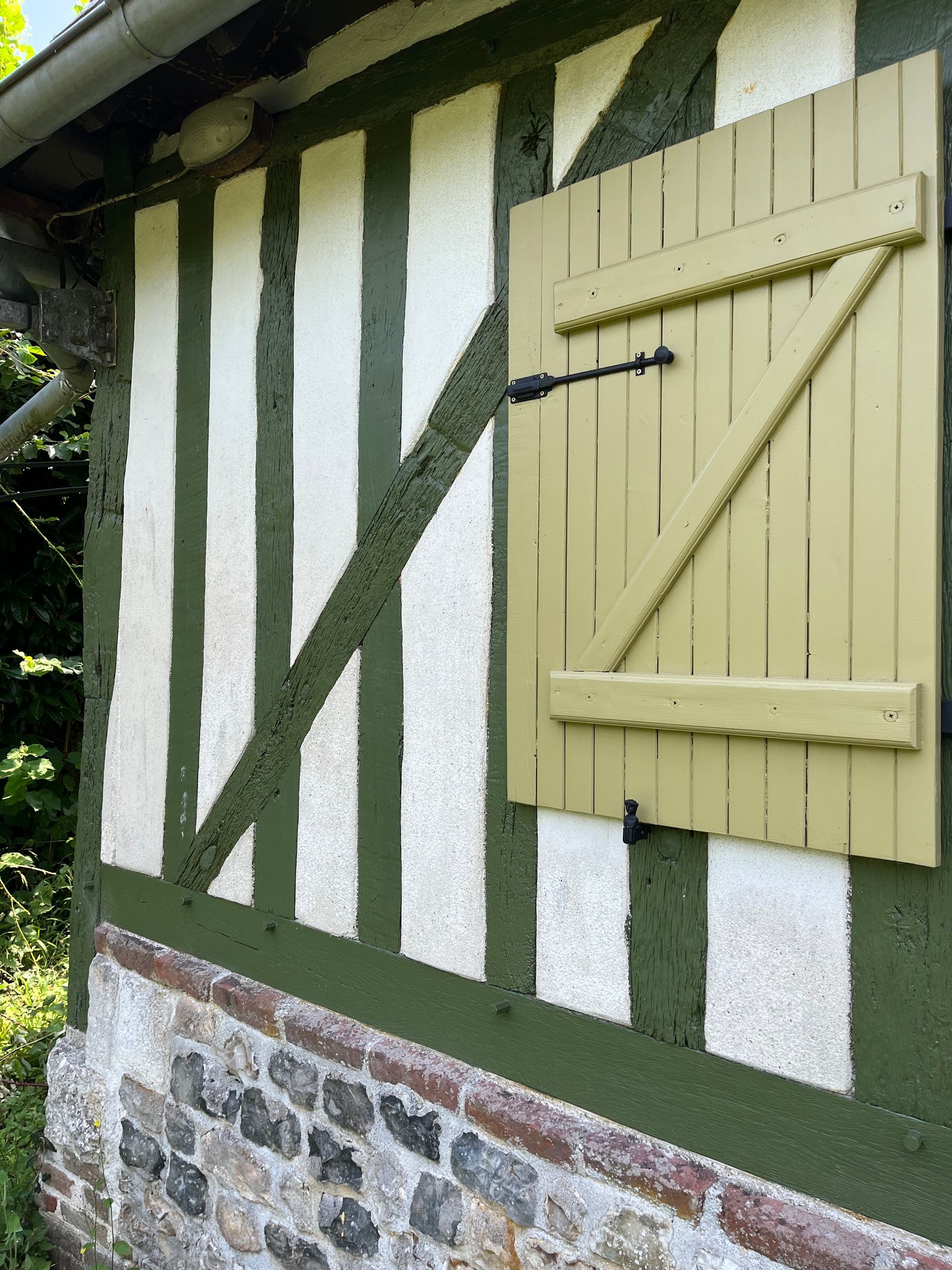 Mur d'un bâtiment à colombages orné de motifs verts et blancs, soubassement en pierre et fenêtre à volets beiges