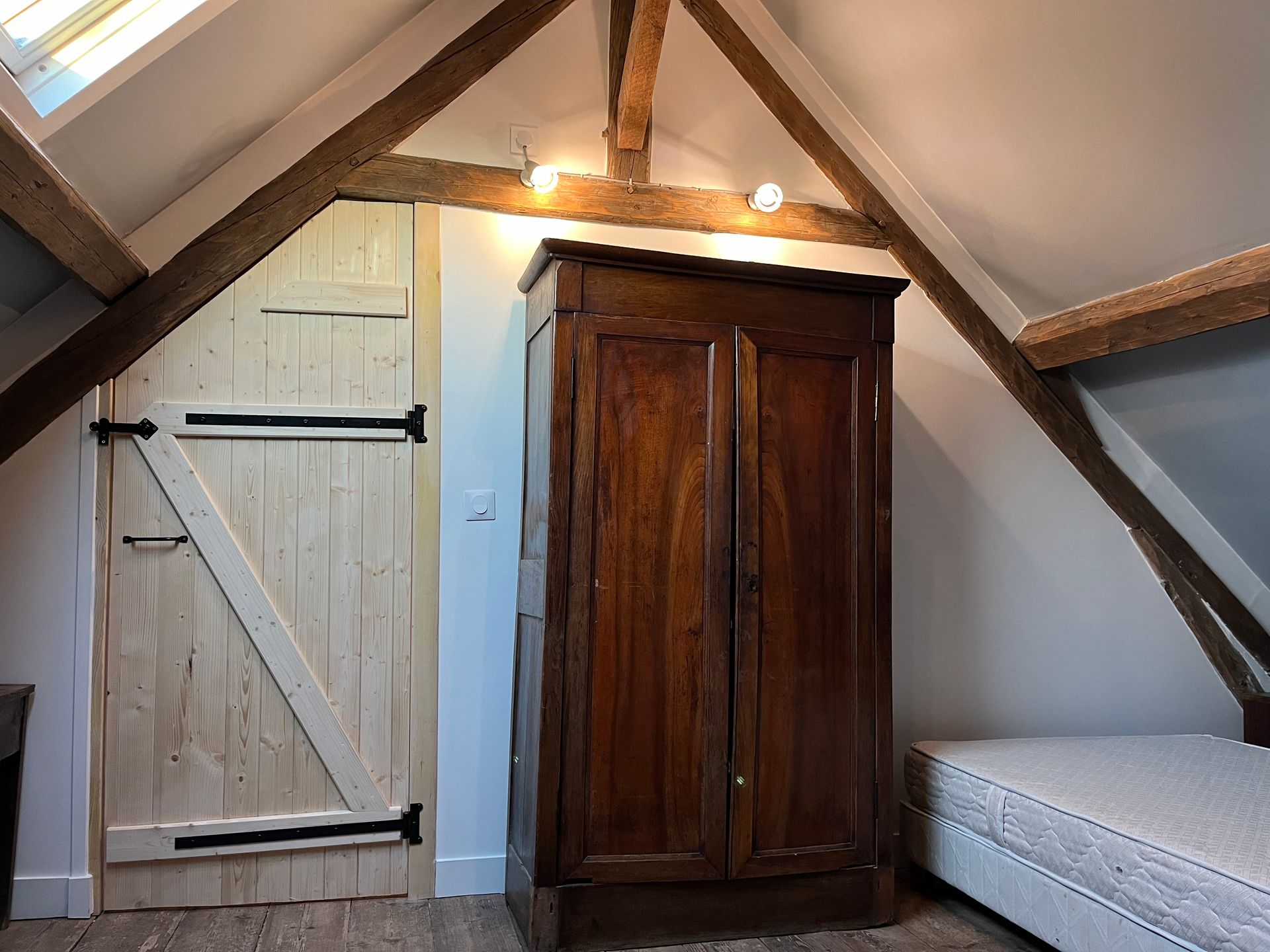 Chambre mansardée avec une armoire en bois, une porte blanche et un lit jumeau sous des poutres inclinées.