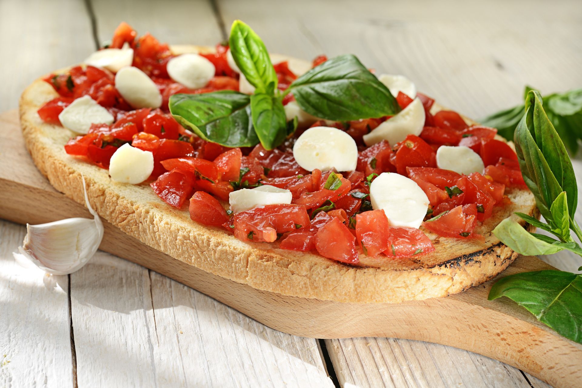Pain grillé garni de tomates en dés, de boules de mozzarella, de basilic, sur une planche de bois.