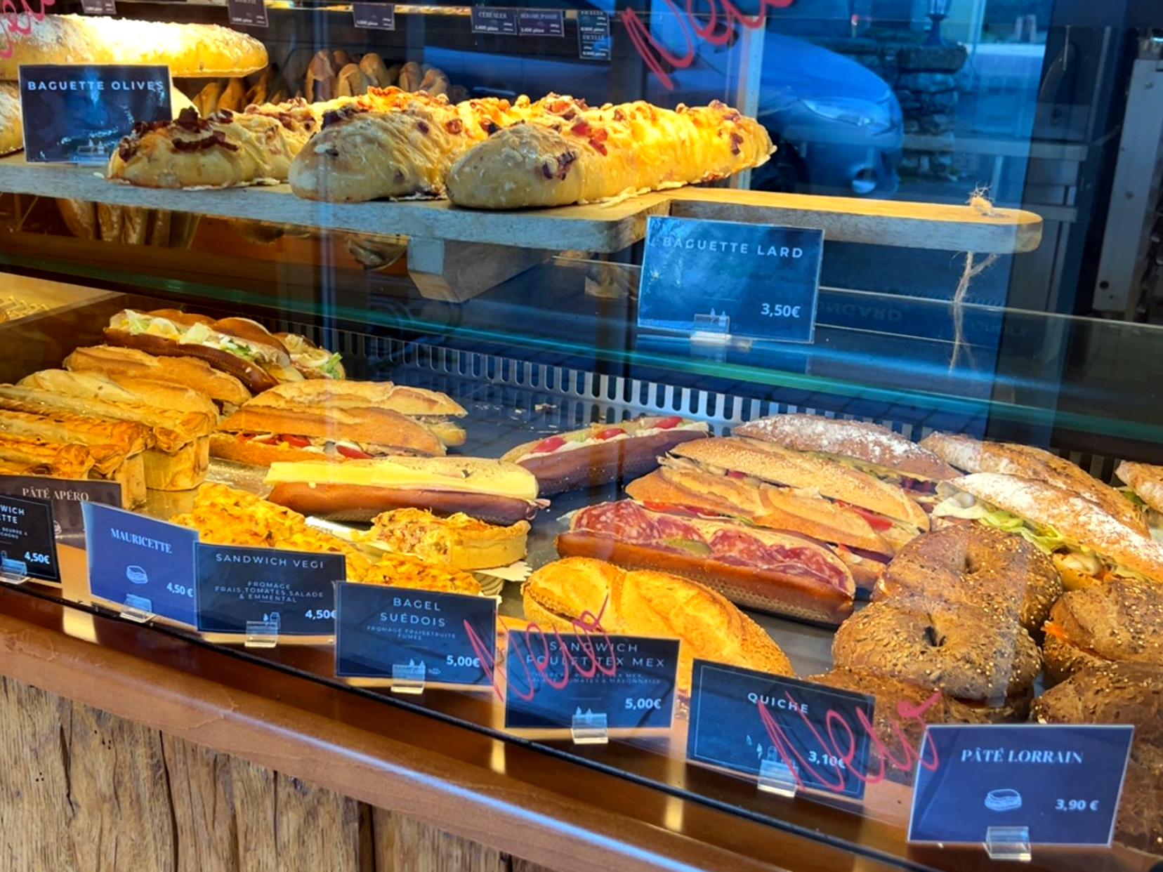 Vitrine de boulangerie avec divers sandwichs et pains, dont baguettes et viennoiseries, accents en bois.