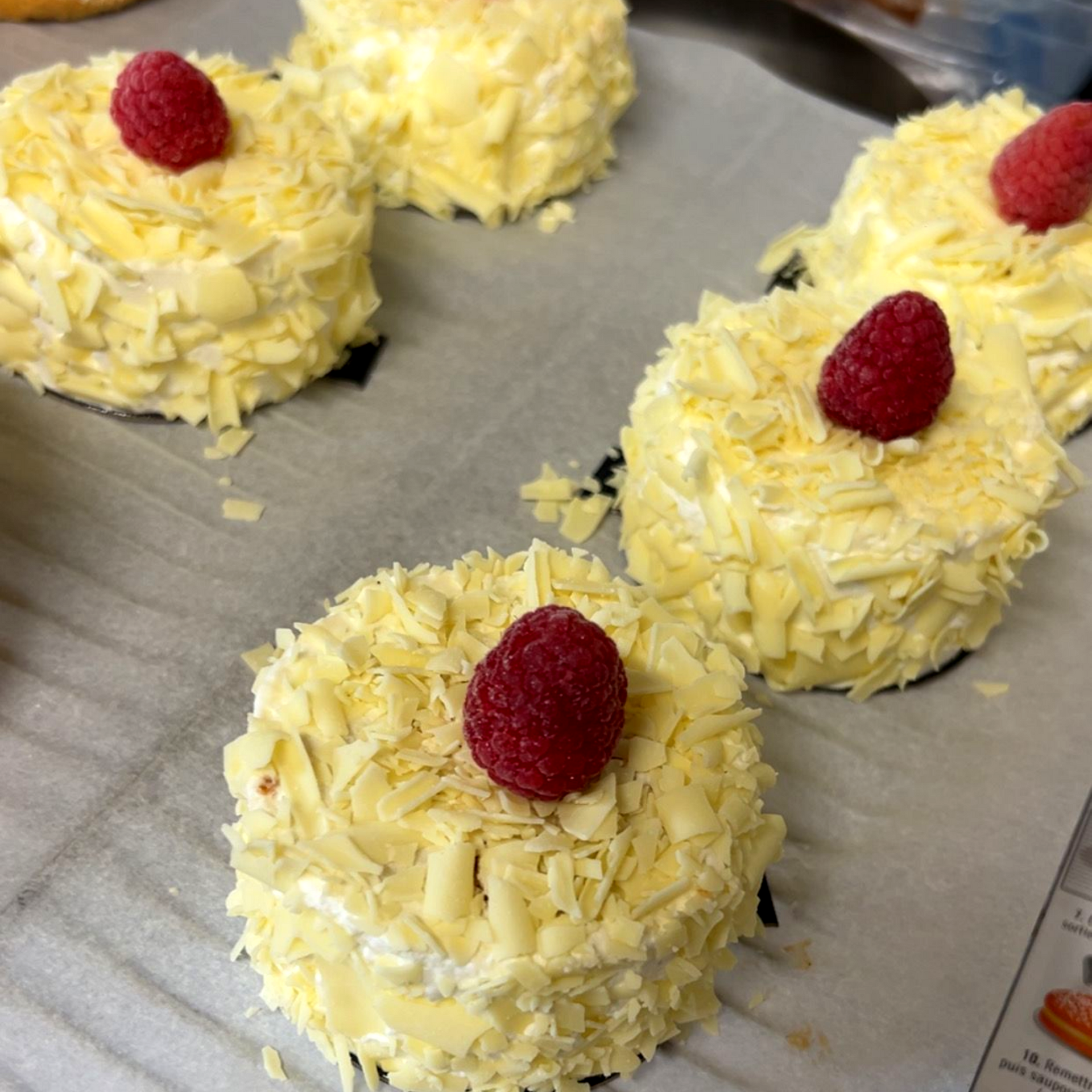 Minigâteaux garnis d'une framboise, enrobés de copeaux de chocolat blanc sur une plaque recouverte de papier sulfurisé.