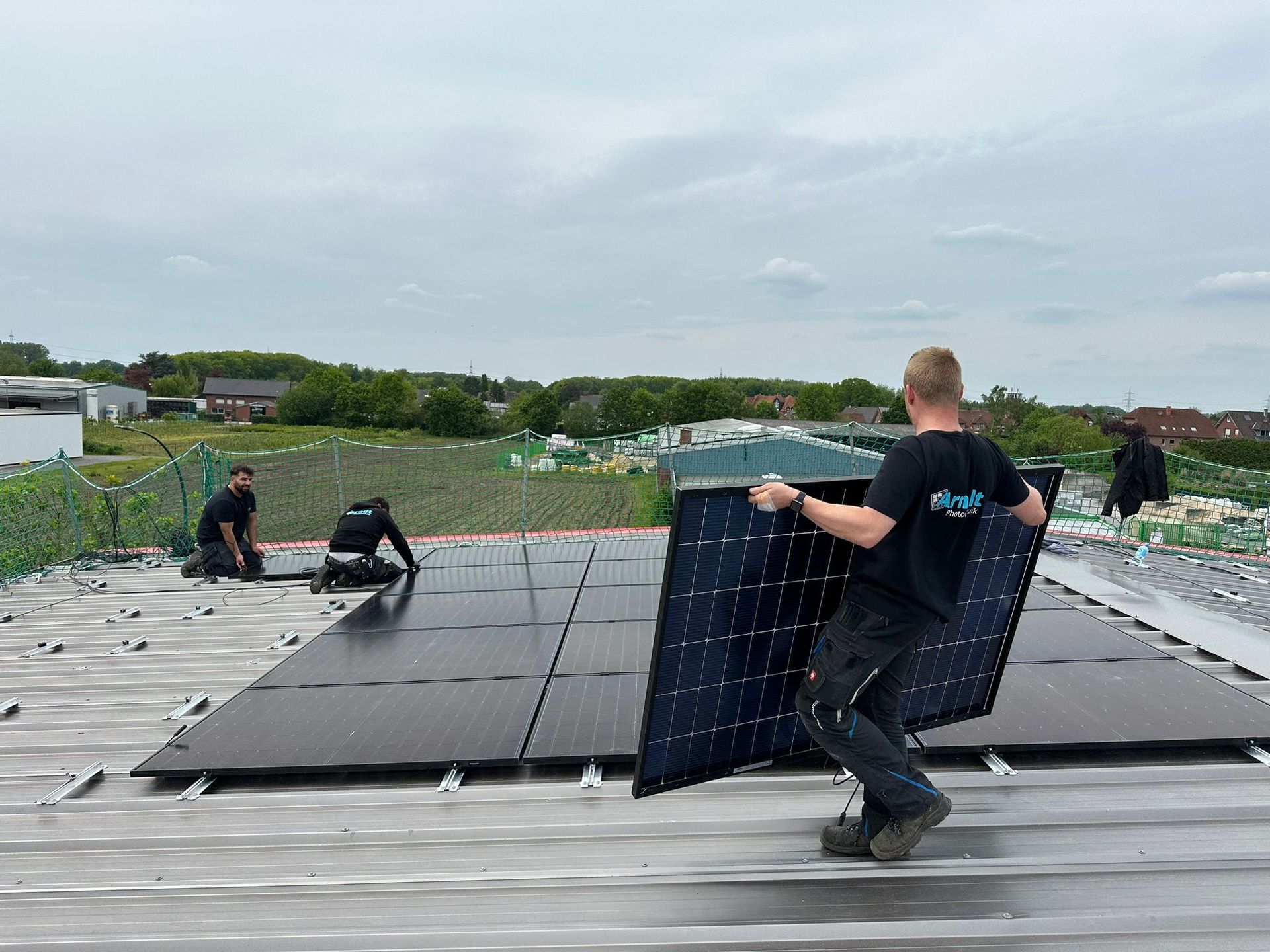 Arbeiter installieren unter bewölktem Himmel Solarmodule auf einem Dach; einer trägt ein Modul.