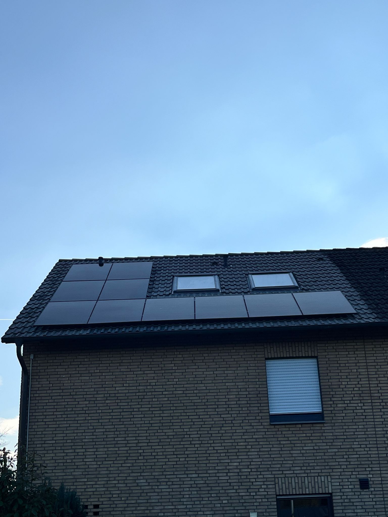 Solarmodule auf dem Dach eines Backsteinhauses mit zwei Oberlichtern vor blauem Himmel.