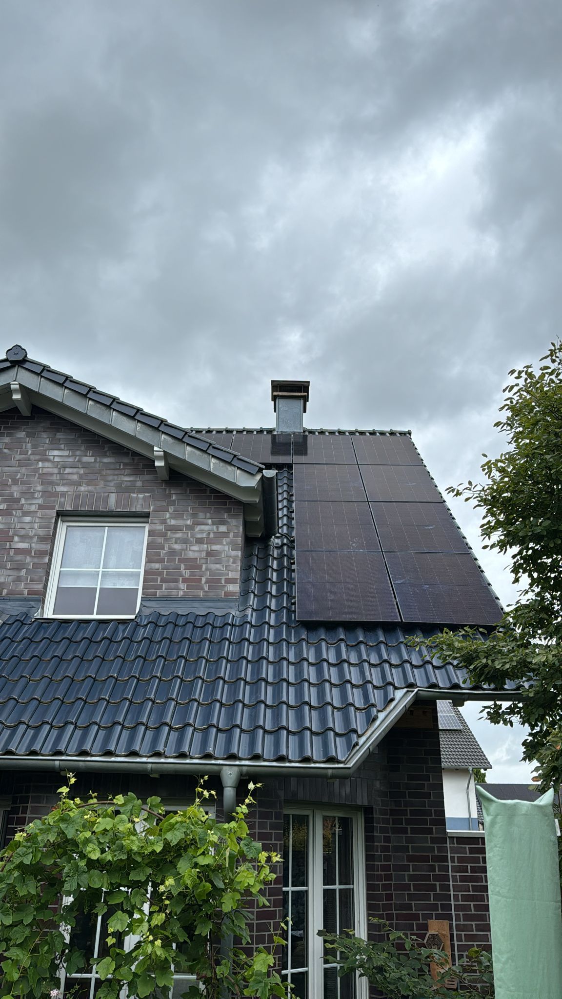 Sonnenkollektoren auf einem dunklen Ziegeldach eines Backsteinhauses unter einem bewölkten Himmel.