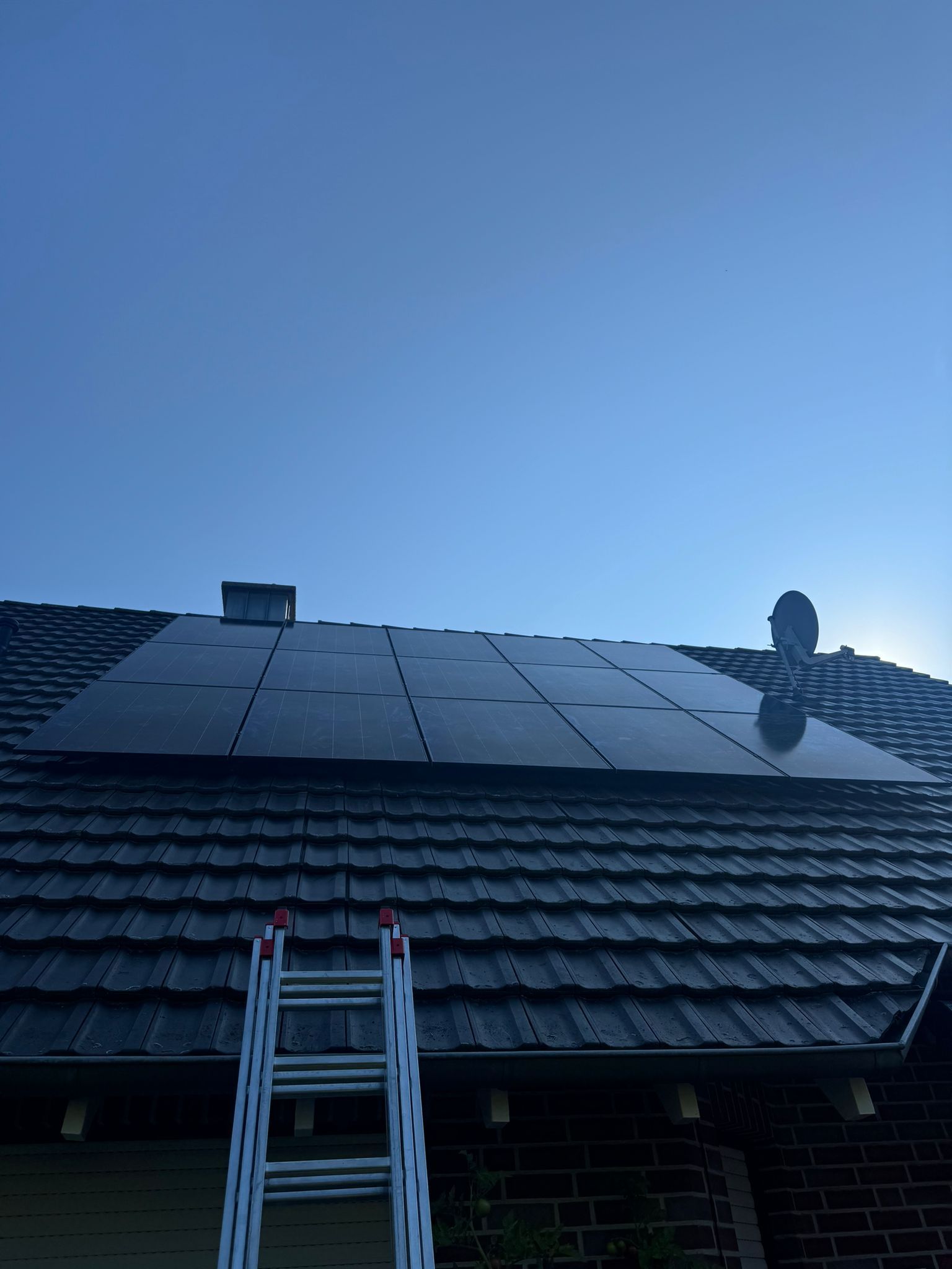 Auf einem dunklen Ziegeldach installierte Solarmodule mit einer Leiter darunter, vor einem blauen Himmel.