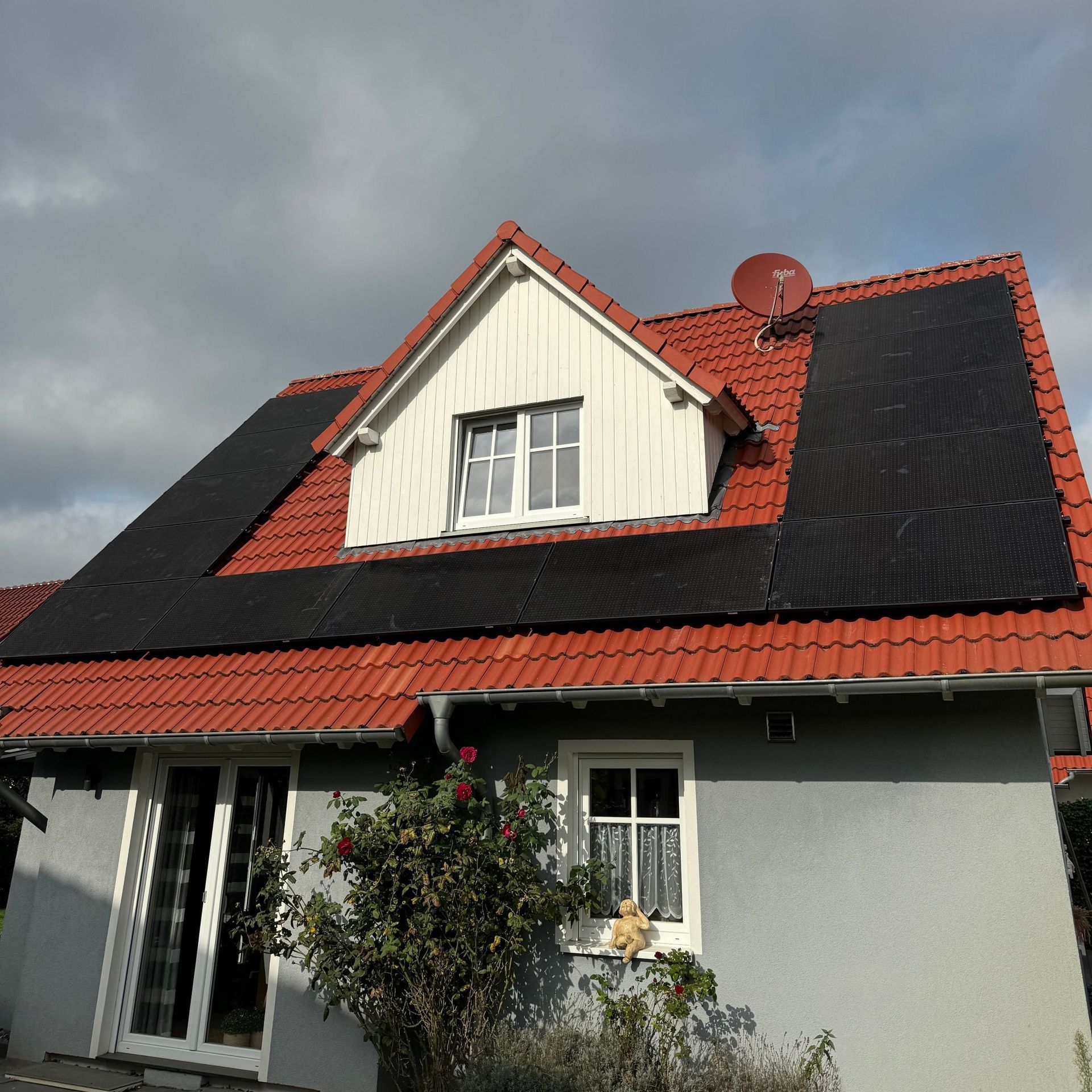 Ein Haus mit rotem Dach, Solarmodulen und einer Satellitenschüssel. Das Gebäude ist grau mit weißen Verzierungen.