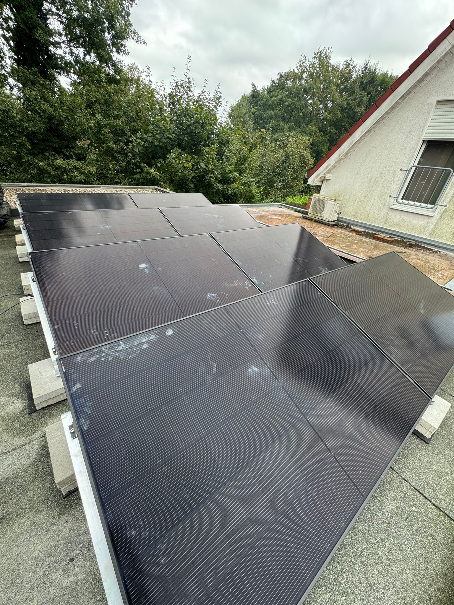 Auf einem Dach installierte Solarmodule, eingebettet zwischen grünen Bäumen und bewölktem Himmel.
