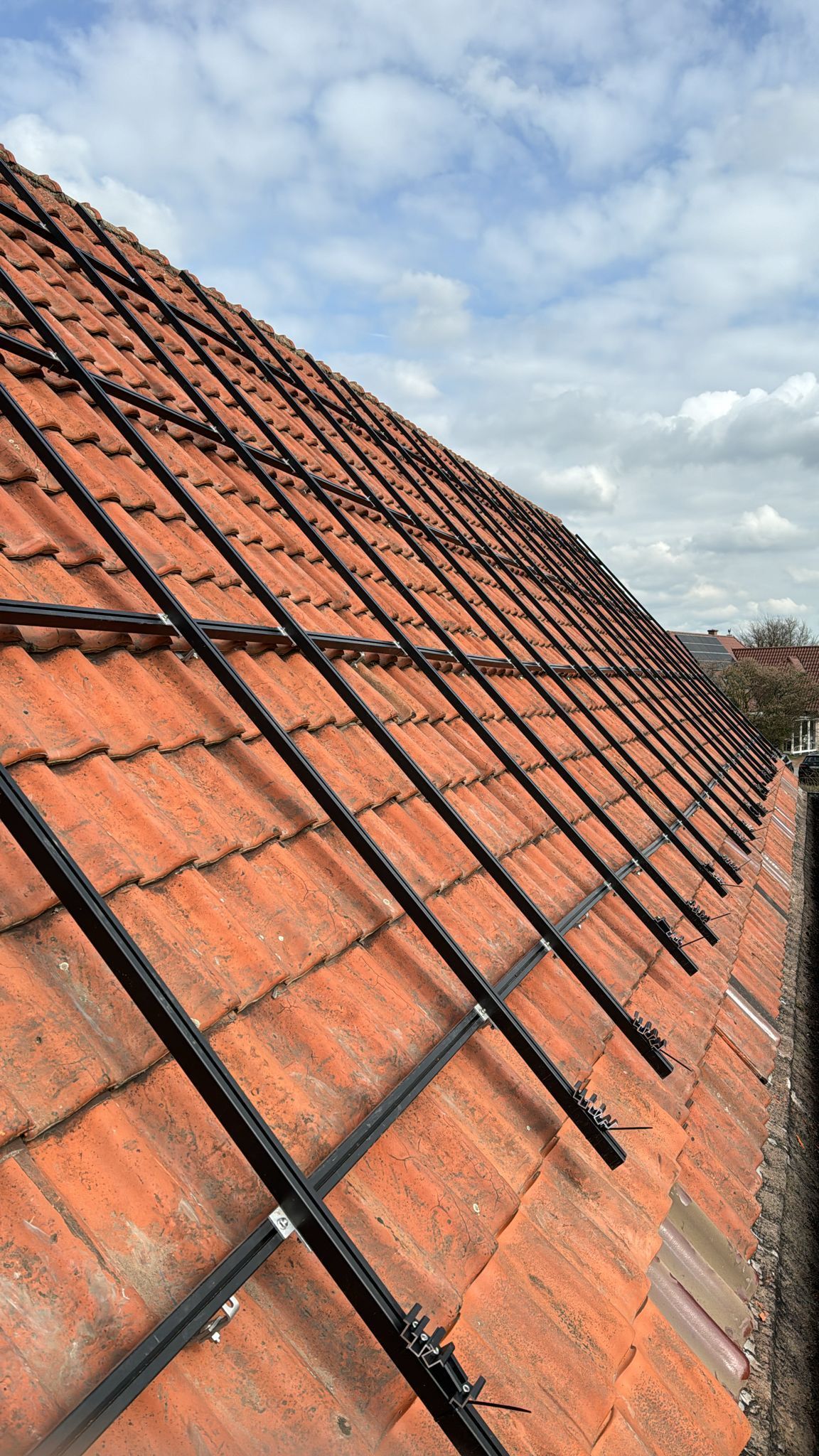 Rotes Ziegeldach mit schwarzen Montageschienen für Solarmodule vor bewölktem Himmel.