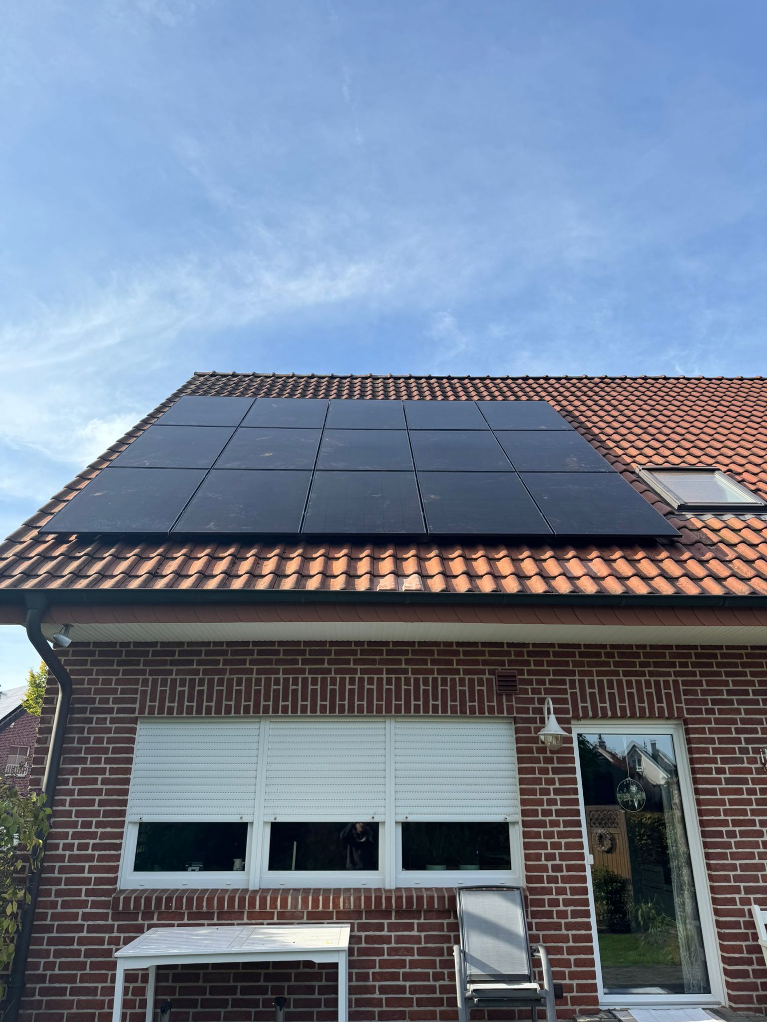 Sonnenkollektoren auf dem roten Ziegeldach eines Backsteinhauses, sonniger Tag. Darunter weiße Jalousien.