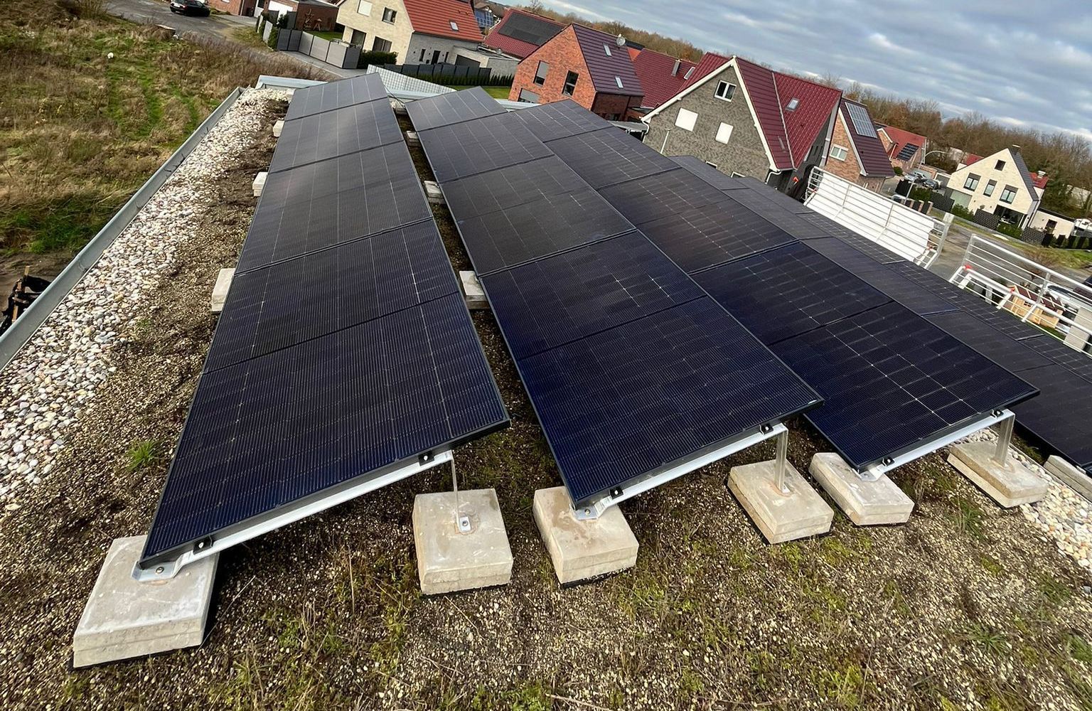 Solarmodule auf einem Flachdach, nach oben gerichtet. Betonblöcke stützen die Module. Grünes Dach mit Gebäuden im Hintergrund.