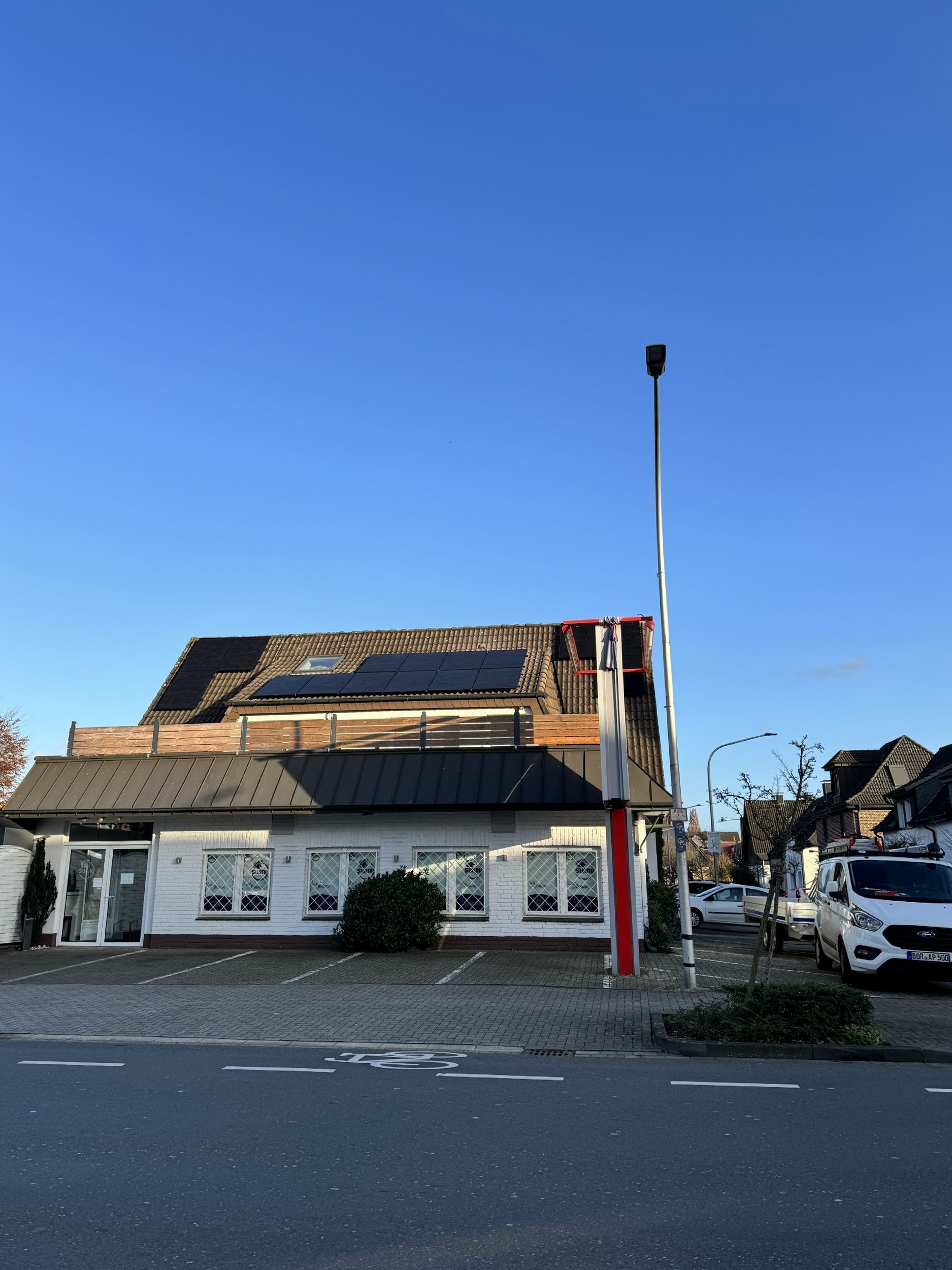 Außenansicht eines weißen Gebäudes mit schwarzem Dach, Solarmodulen und einem rot-weiß gestreiften Mast. Blauer Himmel.