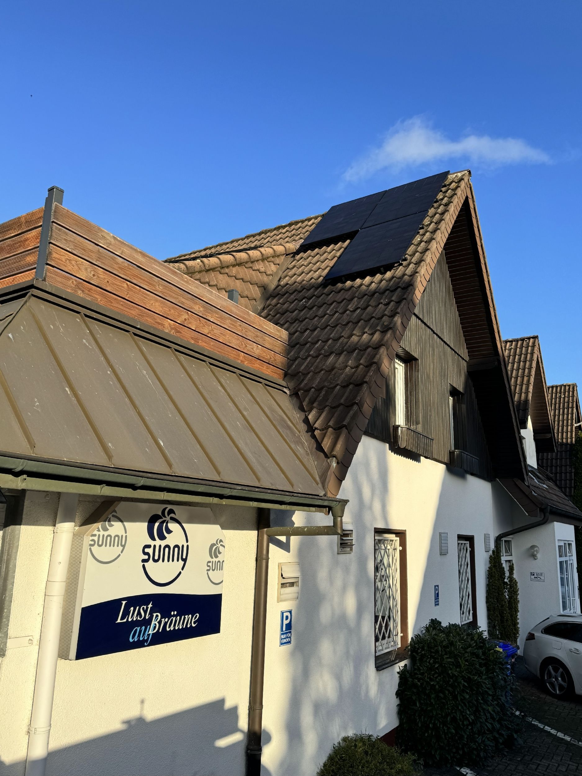 Ein weißes Gebäude mit braunem Dach und Solarmodulen, blaues Schild „Livo Raum“ unter klarem Himmel.