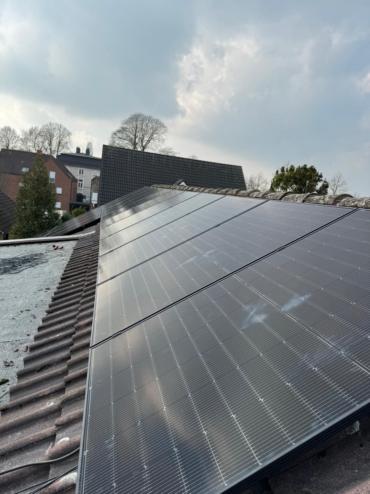 Auf einem Ziegeldach installierte Solarmodule mit Gebäuden und Bäumen im Hintergrund unter einem bewölkten Himmel.