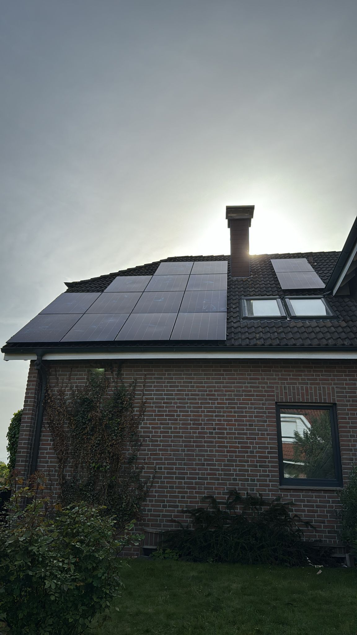 Solarmodule auf dem Dach eines Backsteinhauses, hinter einem Schornstein scheint die Sonne.