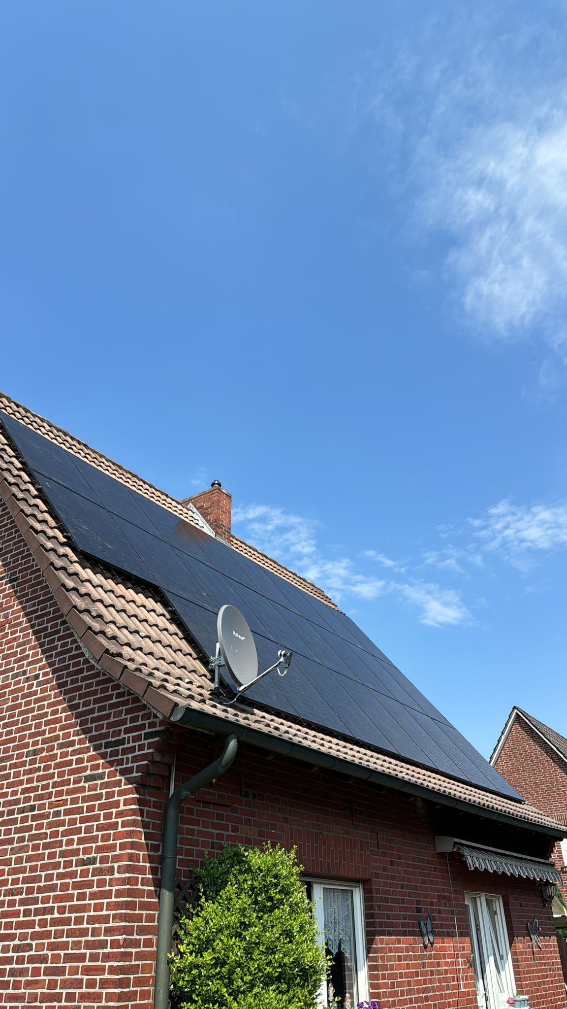 Solarmodule auf dem Dach eines Backsteinhauses mit einer Satellitenschüssel unter blauem Himmel.