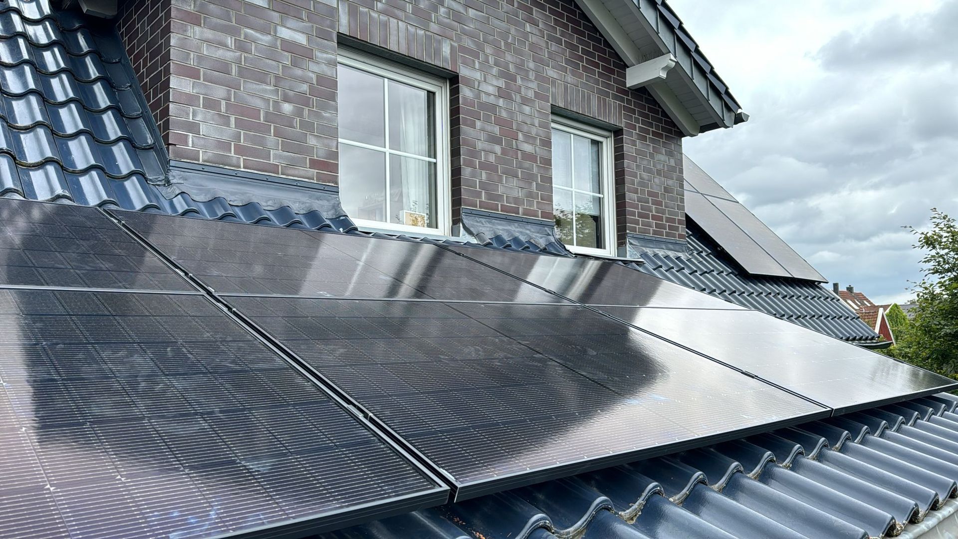 Solarmodule auf einem dunklen Ziegeldach, im Hintergrund ein Backsteingebäude und Fenster.