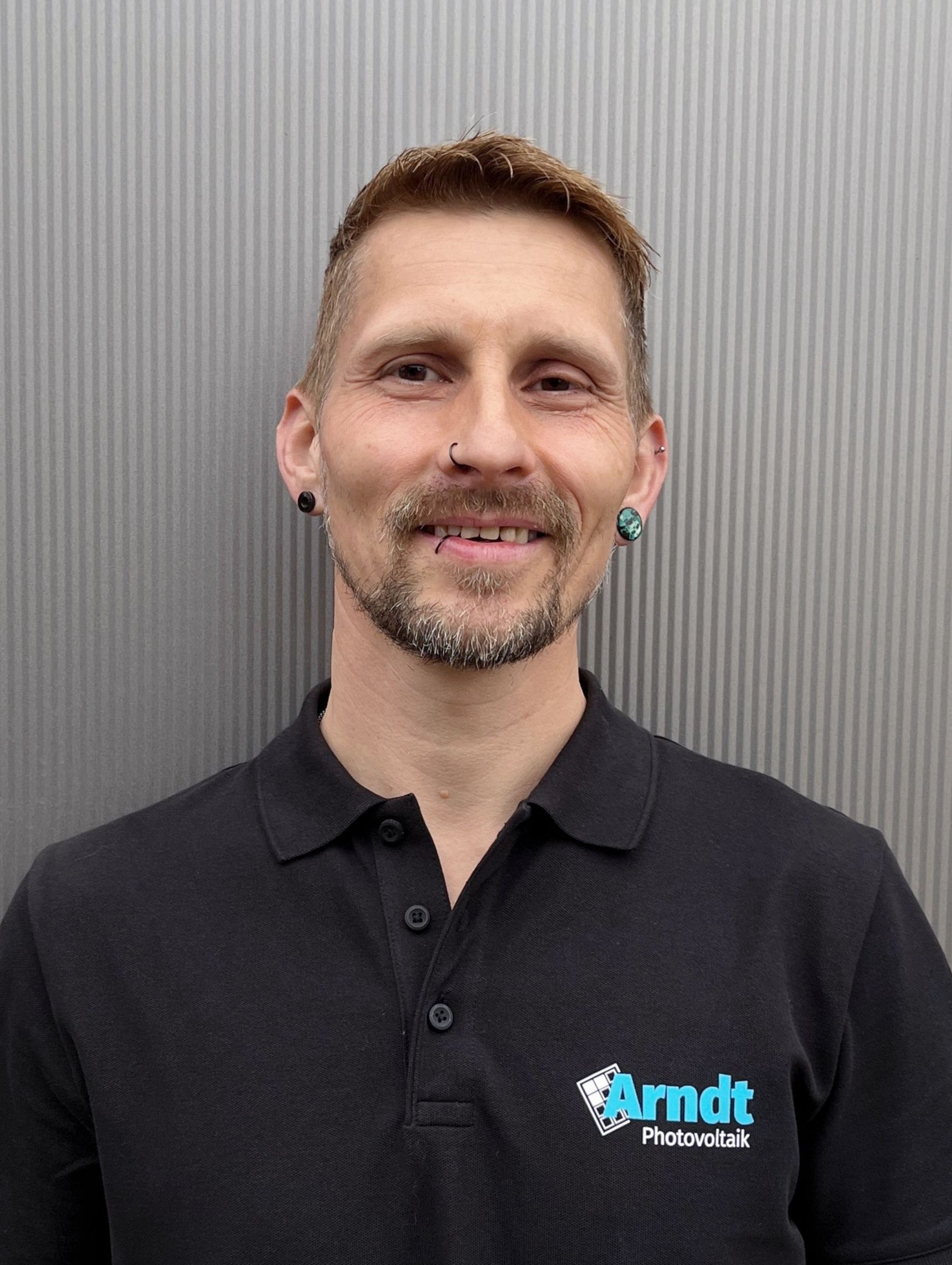 Lächelnder Mann mit kurzen Haaren und schwarzem T-Shirt vor einer Wellblechwand.