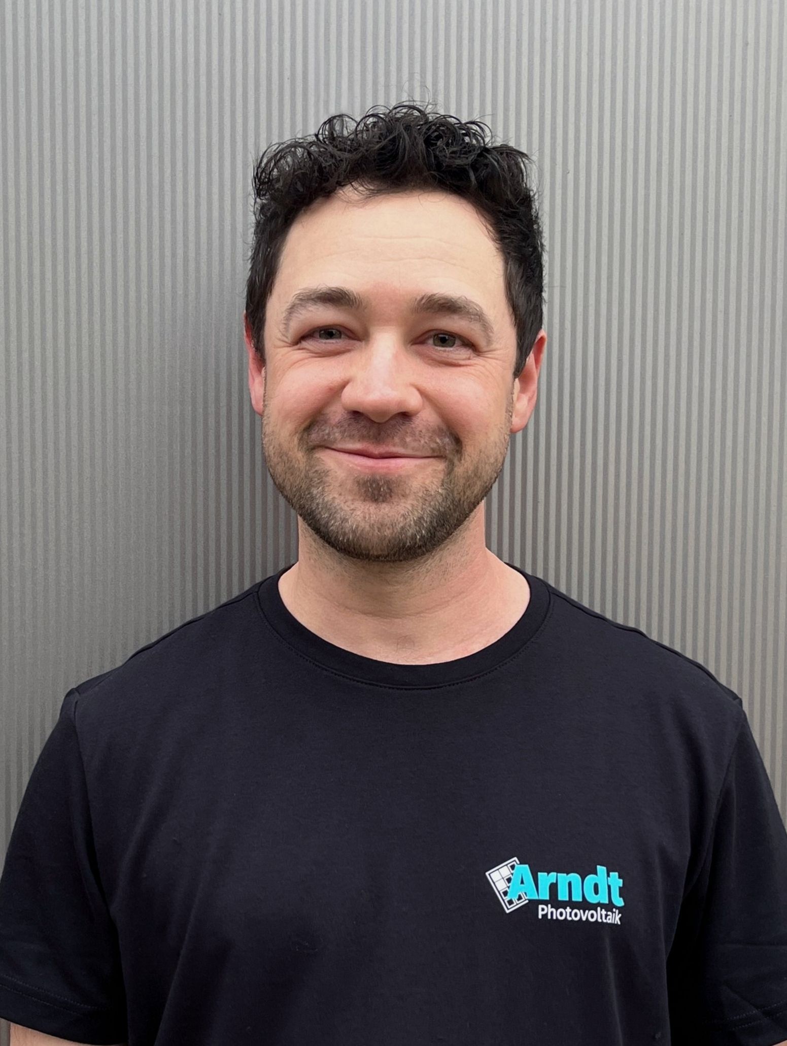 Lächelnder Mann mit dunklen Haaren und schwarzem T-Shirt vor einer Wellblechwand.