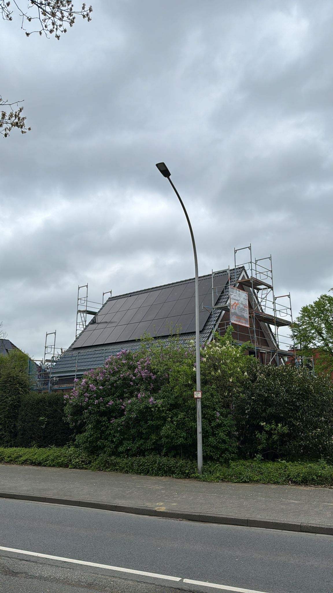 Haus mit dunklen Solarmodulen auf dem Dach, Gerüst und Straßenlaterne unter bewölktem Himmel.