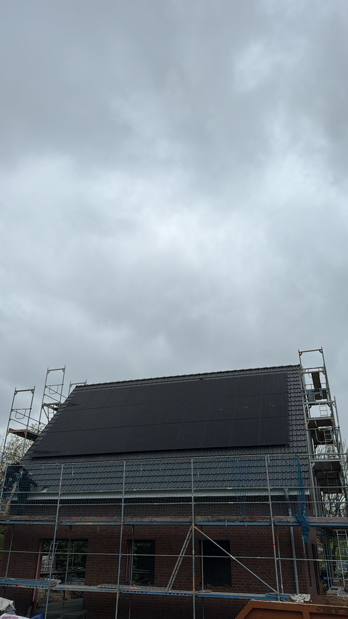 Haus im Bau mit Gerüst und dunklem Dach vor bewölktem Himmel.