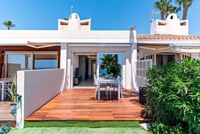 Terraza de madera frente a un edificio blanco con puertas corredizas de vidrio abiertas y mesa exterior.