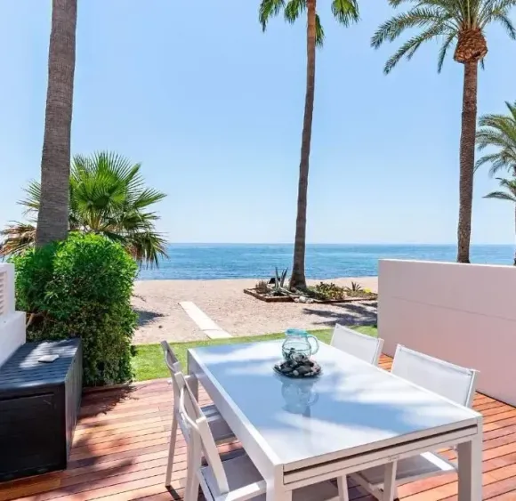 Conjunto de comedor al aire libre en una terraza de madera con vistas a la playa y al océano bajo palmeras.