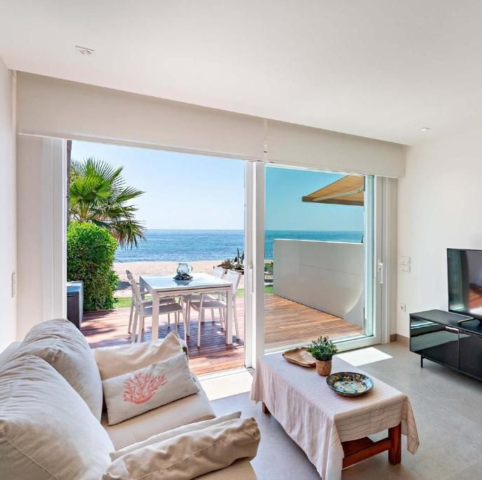 Sala de estar con vista al mar: sofá blanco, puerta corrediza de vidrio, patio con mesa, agua azul.