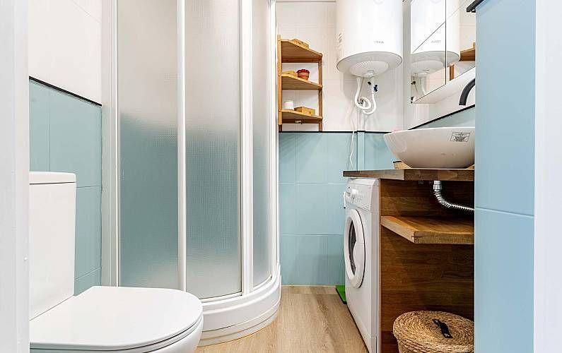 Baño pequeño con ducha, inodoro, lavabo y lavadora. Tonos azul claro y madera.