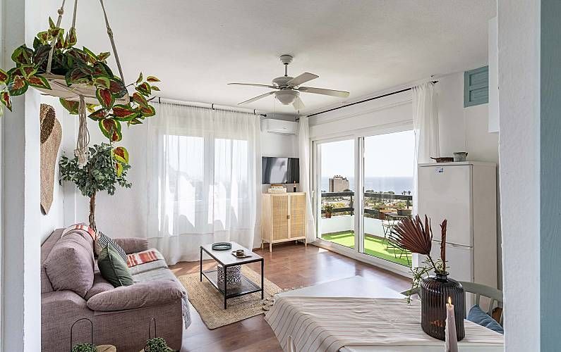 Salón luminoso con plantas, sofá, mesa y vistas al mar a través de una puerta corredera.