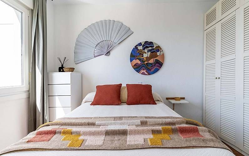 Dormitorio con cama, cómoda, cuadro y armario blanco. Un ventilador decorativo y una obra de arte redonda adornan la pared.