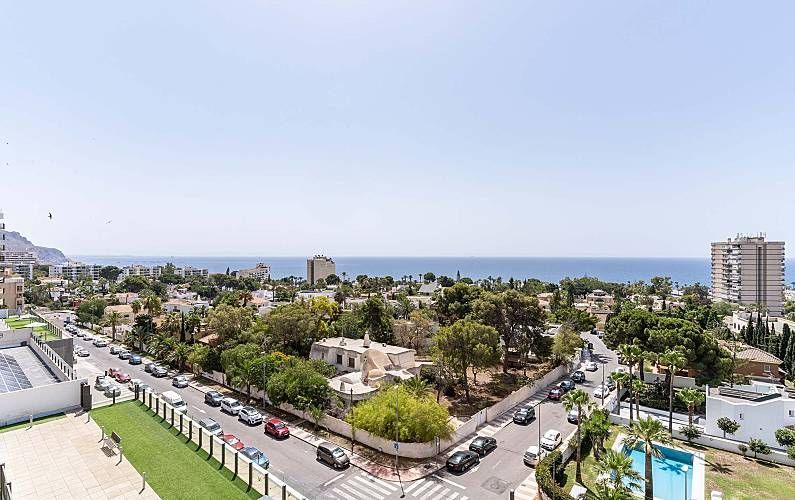 Vista de una ciudad costera con edificios, carreteras, árboles y el océano bajo un cielo azul despejado.