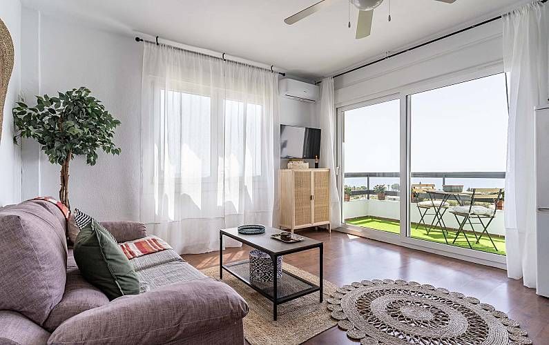 Salón con sofá, televisión y balcón con vistas al mar. Paredes blancas, suelo de madera y alfombra.