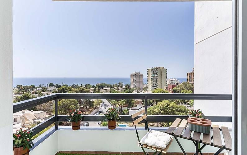 Balcón con vistas al océano, edificios y árboles, con una mesita, plantas y barandilla negra.