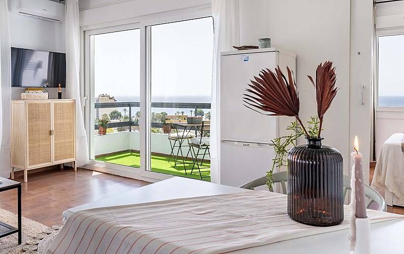 Espacio luminoso con balcón con vistas al mar. Paredes blancas, suelo de madera, jarrón decorativo y armario.