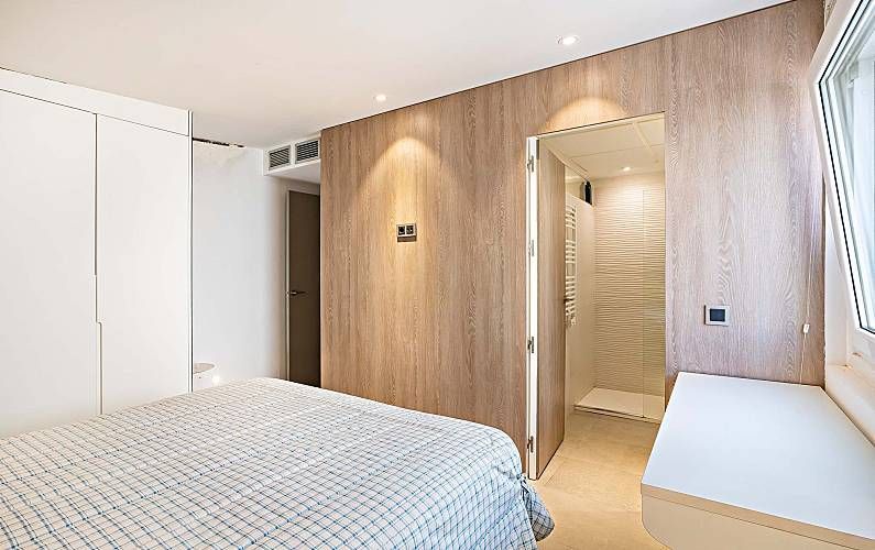 Dormitorio con pared revestida de madera y puerta abierta a la ducha. Cama blanca, armario y ventana.