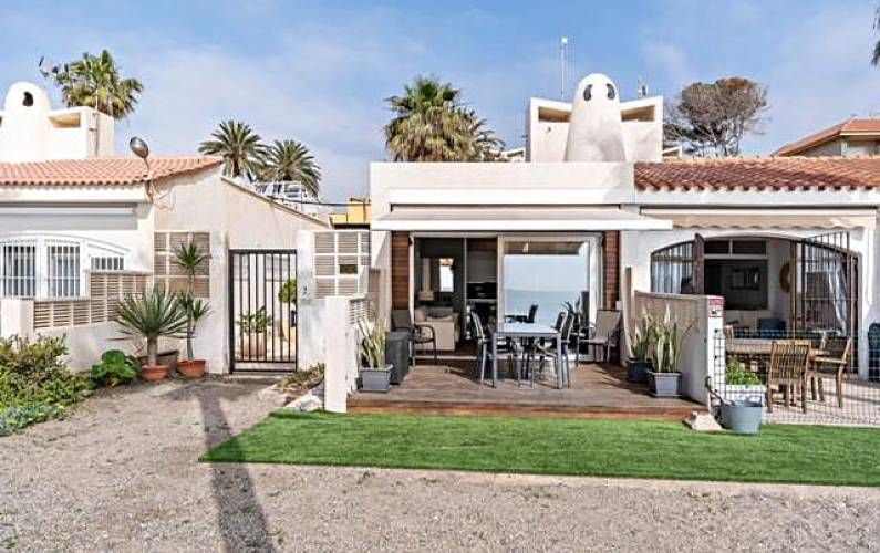 Casas frente al mar con asientos al aire libre, zona de arena y palmeras bajo un cielo brillante.