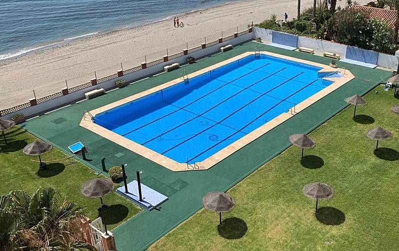 Piscina rectangular cubierta de azul junto a la playa. Se ven el césped, las sombrillas y el océano.